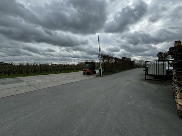 Een geasfalteerde weg met een heftruck en slagboom aan de Graaf van Lynden van Sandenburgweg, omringd door boomgaarden en stapels pallets onder een bewolkte lucht.